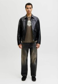 Joven de pie, vestido con una chaqueta de cuero negra, camiseta gráfica marrón, jeans negros desgastados y zapatos negros, contra un fondo claro y liso.