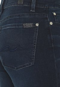 Jeans de ganga azul escuro com um primeiro plano do bolso de trás. Apresenta uma etiqueta e costura em zigue-zague em azul mais claro. Rebites prateados destacados.