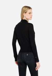 Maglione nero a maniche lunghe con design aderente, texture a coste e bottone dorato di accento sulla parte posteriore del collo. Abbinato a jeans scuri.