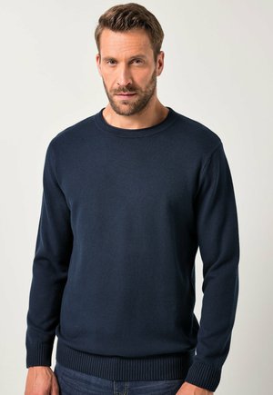 Mann mit kurzen braunen Haaren und Bart, der einen marineblauen Pullover mit langen Ärmeln und dunklen Jeans trägt, vor einem schlichten hellen Hintergrund stehend.