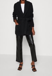 Cardigan nero lavorato a maglia con colletto sciallato, vita con cintura e tasche laterali, abbinato a pantaloni finti in pelle nera a vita alta e sandali neri.