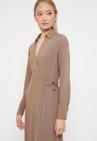 Smeđa haljina na preklop s dugim rukavima, ovratnikom i detaljem vezivanja u struku. Glatka tkanina s suptilnom teksturom, s dubokim V-izrezom.