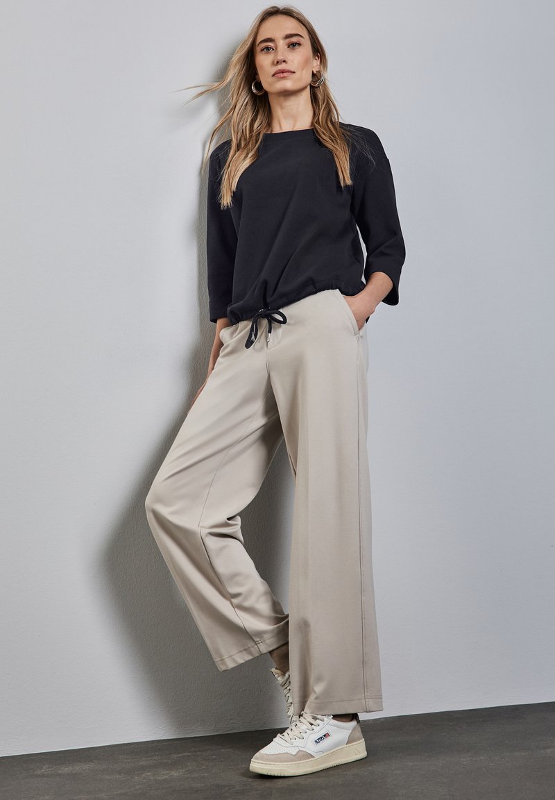 Suéter negro de corte holgado con mangas de tres cuartos, combinado con pantalones anchos beige que tienen un cinturón ajustable. Zapatillas blancas con detalles.