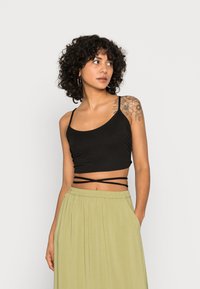 Top cropped preto com alças finas e laços entrelaçados na cintura, combinado com uma saia fluida de verde claro. Textura de tecido suave.