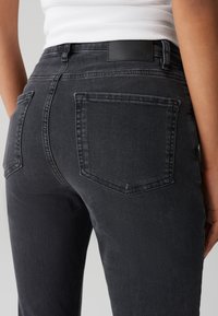 Jeans gris foncé avec une texture lisse, deux poches arrière et une étiquette en cuir sur la taille. La coupe épouse la forme du corps.