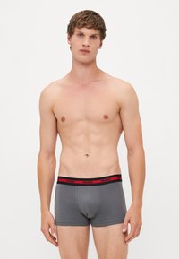 Bóxer ajustado de algodón elástico en gris oscuro con una cinturilla negra que lleva la marca "HUGO" en rojo. Diseño ajustado, textura suave.