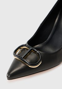 Svart lær pumps med spiss tå, som har en fremtredende sort og gull spennedetalj på vampsiden, med en glatt tekstur og stilrent design.