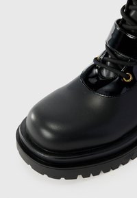Bota de combate negra con punta redondeada, detalles en charol brillante, cordones resistentes y suela de goma texturizada con profundas estrías.