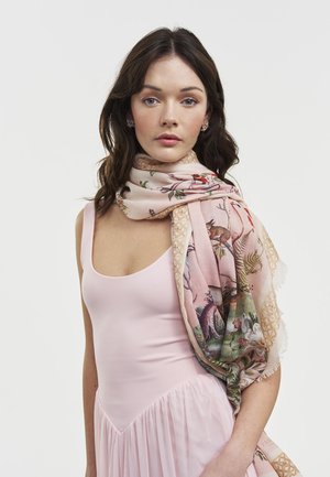 Jauna moteris, tamsiais plaukais, dėvi šviesiai rožinę suknelę ir gėlių rašto šalikėlį, užmėtytą per petį, priešais vienspalvį foną.