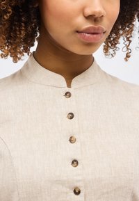 Κοντινή λήψη ενός ατόμου που φοράει ένα μπεζ λινό τοπ με γιακά μανταρίνι και πέντε καφέ κουμπιά στο μπροστινό μέρος.