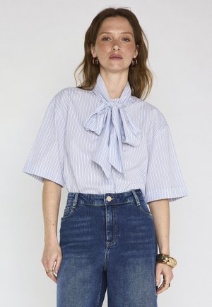 Donna che indossa una blusa a maniche corte a righe azzurre con un grande fiocco al collo, jeans blu a vita alta, orecchini a cerchio dorati e un braccialetto spesso oro e nero.