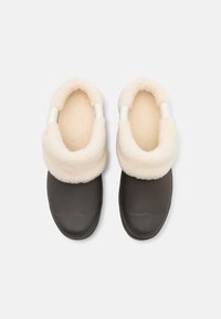 Mörkbruna träskor med en mjuk, off-white fleecefoder och en vikbar krage. Det övre materialet ser slätt ut och är något glansigt.