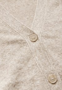 Cardigan beige con scollo a V, lavorazione a maglia testurizzata e un singolo bottone tondo di colore chiaro sul davanti.