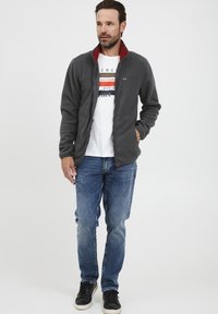 Grå fleecejakke med rød krage, glidelås foran, kombinert med en hvit grafisk t-skjorte og blå jeans, båret med svarte joggesko.