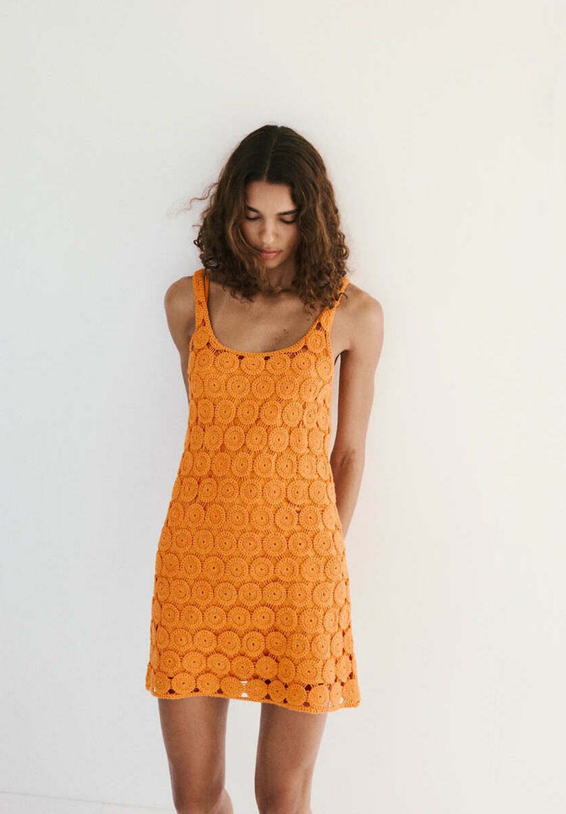 Femme aux cheveux bouclés portant une robe orange en crochet sans manches, debout contre un mur blanc, regardant vers le bas les mains derrière le dos.