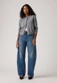 Cardigan gris en tricot avec un col rond et une fermeture à boutons, associé à un jean bleu à jambe large. Des chaussures noires à bout pointu complètent le look.