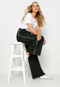 Pantalons larges noirs avec des rayures blanches sur le côté, associés à un haut blanc à manches courtes et des baskets blanches, assis sur un tabouret blanc.