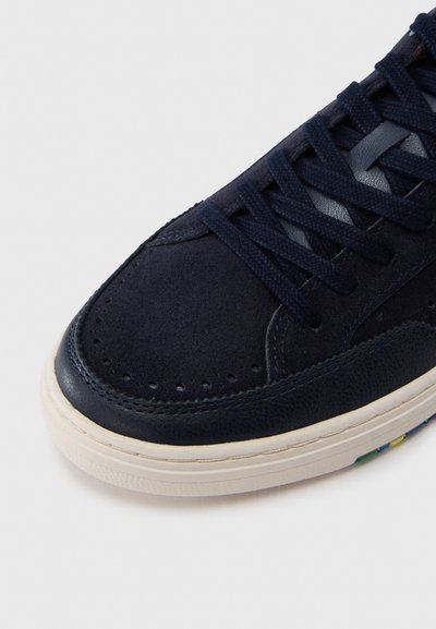 Ακμή αθλητικού παπουτσιού σε ναυτικό μπλε με υλικά σουέντ και δέρματος, διάτρητες λεπτομέρειες, ναυτικά κορδόνια και λαστιχένια σόλα σε εκρού χρώμα με πολύχρωμες πινελιές.