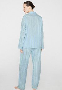 Mujer de pie descalza, vistiendo un top de manga larga con rayas azul claro y pantalones a juego, de espaldas contra un fondo blanco liso.