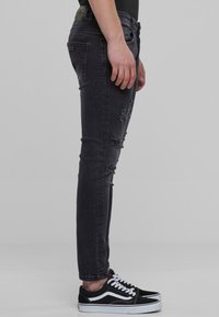 Jeans ajustados negros con detalles desgastados, presentando un clásico diseño de cinco bolsillos, combinados con zapatillas negras y blancas.