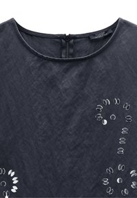 Czarna bluzka o okrągłym dekolcie i zapięciu na zamek z tyłu. Posiada dekoracyjne zdobienia w kształcie okrągłym i owalnym.