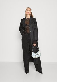 Γυναίκα ντυμένη με μαύρο μακρύ παλτό, διαφανή κεντημένη μπλούζα, μαύρα τζιν, μαύρες μυτερές γόβες, κρατώντας μια μικρή μεταλλική τσάντα μπροστά σε απλή φόντο.