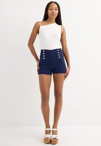 Bodysuit blanco de un solo hombro combinado con pantalones cortos de talle alto en color navy con botones decorativos. La modelo lleva sandalias de plataforma blancas.