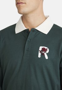 Dark green polo shirt with a white collar. Features an embroidered red rose accent and a white letter "R" on the left side.