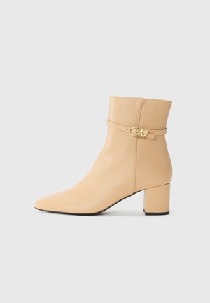 Beige leren enkelboot met een puntige neus, chunky hak en een decoratieve enkelriem met een gouden gespdetail.
