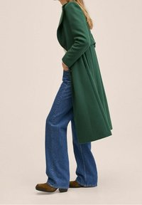 Lang grøn frakke med skræddersyet pasform og plissédetalje, kombineret med blå jeans med vide ben og brune støvler. Glat stoftekstur.