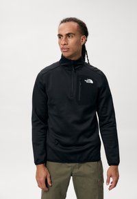 Sudadera de forro polar negro con cremallera, cuello alto, bolsillo frontal en el pecho y el logo de The North Face. Textura suave, diseño ajustado, mangas largas.