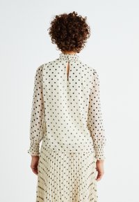 Blusa crema con pois neri, caratterizzata da un collo alto, una apertura a chiave con bottone e polsini arricciati. Maniche lunghe e tessuto leggero.