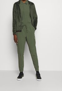 Mikina na zip z fleecu a odpovídající tepláky v tmavě zelené barvě. Mikina má límec; tepláky mají elastický pás a boční kapsy. Černé tenisky.