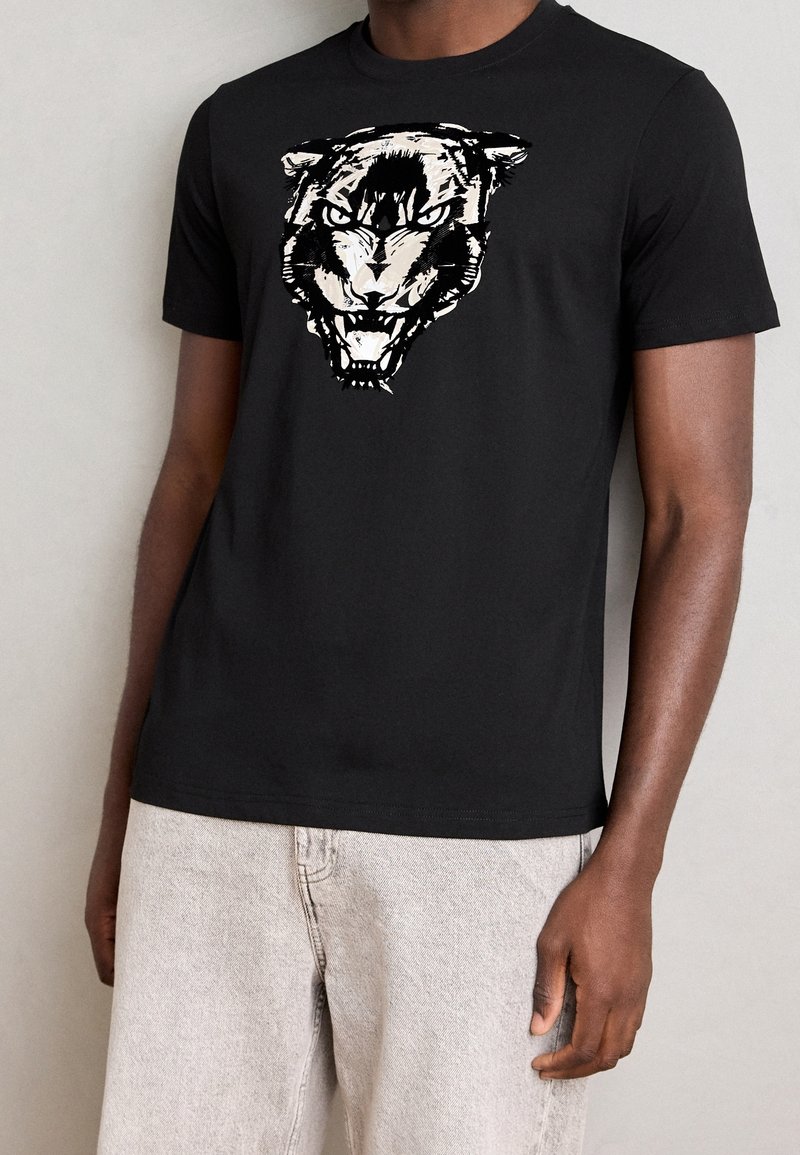 Černé bavlněné tričko s grafickým motivem stylizované tygří hlavy v bílé barvě. Krátké rukávy s kulatým výstřihem, hladká textura.