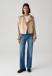 Giacca corta beige con ampio colletto e bottoni doppi, abbinata a jeans a gamba larga blu e scarpe nere. Texture liscia.