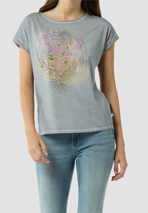 Femme portant un t-shirt gris clair à manches courtes, avec un motif floral et géométrique jaune et rose, associé à un jean bleu clair sur un fond uni.