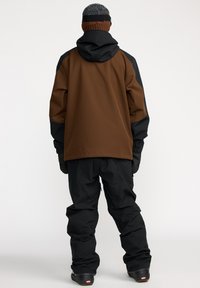 Ski-jack en -broek in bruin en zwart, met een capuchon, gestructureerde stof en elastische manchetten, ontworpen voor winteractiviteiten.