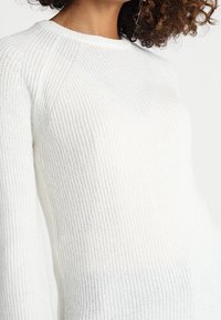 Valkoinen neulepaita, jossa on ribbipinta, pyöreä kaula-aukko ja pudotetut olkapäät. Hihamme ovat pitkät ja malli on slim fit.