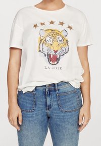 T-shirt bianco a maniche corte con una stampa di un volto di tigre, decorato con stelle dorate e marroni sopra e il testo "LA JOIE" sotto.
