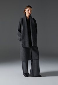 Abrigo de lana gris con un corte holgado, botones grandes y una bufanda a juego. Combinado con pantalones grises de pierna ancha y zapatos negros. Diseño simple.