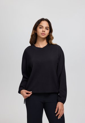 Junge Frau mit welligem Haar, die ein schwarzes Langarmshirt und schwarze Hosen trägt, steht vor einem schlichten hellgrauen Hintergrund.