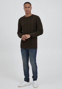Pulover tricotat de culoare maro închis, cu un model în diamant și detalii cu cablu, asortat cu blugi albaștri și adidași albi. Modelul își ține mâinile laolaltă.