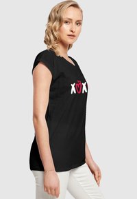 Svart t-skjorte med korte ermer som har et grafisk design med røde og hvite "XOXO"-bokstaver. Stoffet virker glatt og lett.