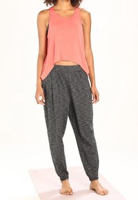 Frau, die barfuß auf einer Yogamatte steht und ein lockeres, ärmelloses Oberteil in Korallenfarbe sowie eine dunkelgraue, lässige Hose trägt.