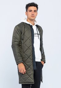 Gequilteter, olivgrüner langer Mantel mit Reißverschluss, darunter ein weißer Kapuzenpullover und schwarze Hose. Glatte Textur und modernes Design.