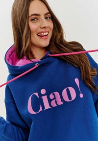 Modrá mikina s růžovým vnitřkem a šňůrkami, s textem "Ciao!" v tučném růžovém písmu na přední straně. Měkká textilie.