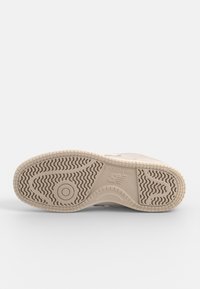 Semelle beige de sneaker New Balance montrant des motifs de la bande de roulement, rainures circulaires et ondulées, avec le logo New Balance embossé au centre.