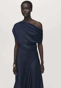 Navy blue, draped off-the-shoulder dress made of soft fabric with a fitted waist and wide flowing skirt. Silver bangle on the wrist.