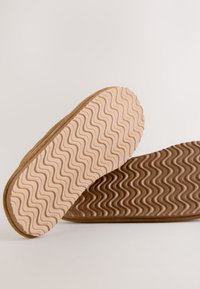 Miękkie zamszowe buty wsuwane z beżową, teksturowaną podeszwą o falistym wzorze. Podeszwa jest jasnobrązowa, co stanowi kontrast dla cholewki.