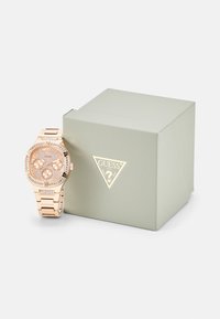 Montre-bracelet en or rose avec un cadran rond, un visage texturé rose, des accents en cristal et un bracelet en métal assorti, accompagnée d'une boîte cadeau vert clair.